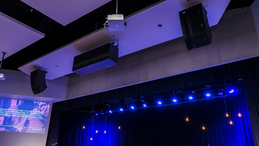 A modern church with a projector and speakers mounted on the ceiling and purple stage lights.